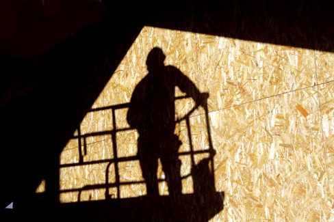 L'ombre d'un homme casqué debout sur une nacelle se reflète sur une plaque d'OSB lors d'un suivi photographique de chantier.
