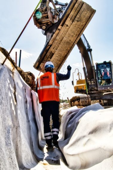 Dans cette photographie de suivi de chantier, un ouvrier casquée guide par ses gestes un tracts-elle qui déverse dans une tranchée un lit de graviers lors du chantier de réfection de la RN 165 à la sortie de Nantes en direction de Vannes