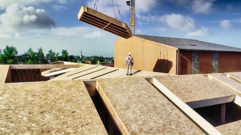 Un charpentier casqué et muni de son harnais, attend seul sur une toiture de type OSB qu'un panneau de bois soit approvisionné par une grue de type PPM à tour.