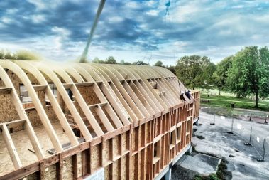 Deux compagnons charpentiers casqués attendent sur une charpente que la grue approvisionne d'autres éléments de toiture.