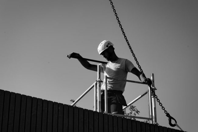 Un charpentier sur une toiture tient une masse en équilibre sur son épaule. Encore un reportage photo et suivi de chantier réussis pour Olao