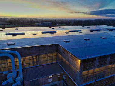 Lever de soleil sur une toiture. L'étanchéité quasi neuve reflète les premiers rayons bleus et jaunes de ce lever de soleil sur l'usine CLT du groupe Piveteau bois à Sainte Florence, aux Essarts en Bocage (85). Les lumières sont déjà allumées dans l'usine. Les machines tournent à plein pour fabriquer des pièces de bois sur mesure. C'est aussi le site de fabrication des murs, des panneaux en CLT. Cet acronyme signifie Cross Laminated Timber, ou X-Lam en anglais. En même temps, une autre machine est livrée. La demande est forte. Nous allons filmer pendant toute la journée de cette presque chaude journée de décembre.