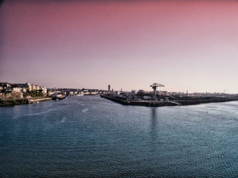 Depuis la pointe de Trentemoult, dans un ciel rose pâle, la grue Titan grise se dessine au loin. La Loire se divise en deux bras distincts sous l'oeil de la grue.