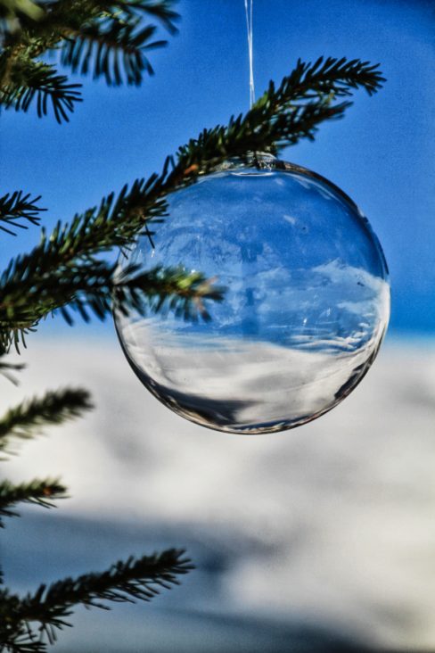 Une boule de Noël translucide est accrochée à une branche de sapin à l'extérieur. La montagne et la neige se reflète dans ce qui semble être une petite planète bleue et nuageuse.
