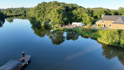 Une toue cabanée sur la Mayenne se prépare à apponter. Un homme est debout sur la proue. Il est coiffé d'un canotier et tend vers le rivage une coupe de champagne dans chaque main.