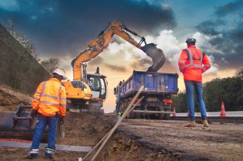 Un conducteur de travaux de chez NGE contrôle les travaux d'excavation d'une pelle. Un camion-benne bleu est à côté de la pelle et reçoit de la terre. Un ouvrier est dans la tranchée et vérifie si le niveau est bon.