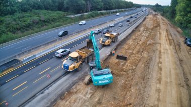 Une pelle de couleur bleu ciel est en train d'alimenter un chargeur positionné à ses côtés. D'énormes pierres sont arrachées du sol avant d'être chargées. Un autre chargeur aux couleurs également de NGE est placé juste derrière en attente, afin de ne pas casser le rythme.