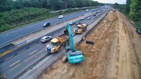 Une pelle de couleur bleu ciel est en train d'alimenter un chargeur positionné à ses côtés. D'énormes pierres sont arrachées du sol avant d'être chargées. Un autre chargeur aux couleurs également de NGE est placé juste derrière en attente, afin de ne pas casser le rythme.