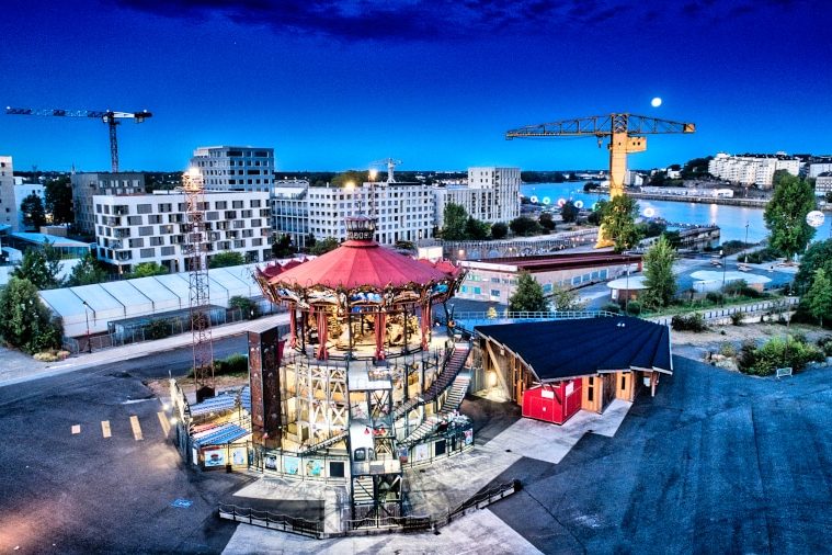 Le carrousel des Mondes marins imaginé par les célèbres Machines de l'île brille de mille feux à l'heure bleue. Les anneaux de Buren dans le fond tente d'attirer la pleine lune qui tutoie la grue jaune
