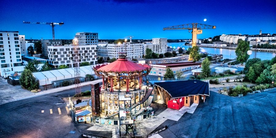 Le carrousel des Mondes marins imaginé par les célèbres Machines de l'île brille de mille feux à l'heure bleue. Les anneaux de Buren dans le fond tente d'attirer la pleine lune qui tutoie la grue jaune