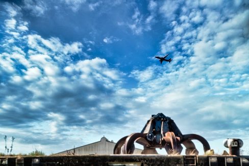 une griffe de type industrielle servant au déchargement des bateaux est posé sur un ponton de béton. La photographie est prise depuis la Loire sur une embarcation. Dans le fond, un ciel bleu chargé de cumulus. Un avion décollant de l'aéroport de Nantes Atlantique vient déchirer le ciel. Le monde d'après, où le passage d'une avion devient presque un miracle. La fin d'une époque ?