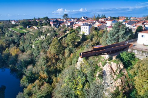 Photographie aérienne, au dessus de la Maine. Sur cette image, un homme masquée s'est avancé sur le nouveau belvédère le "Porte-Vue" imaginé par Emmanuel Ritz et s'inscrivant dans le cadre du Voyage à Nantes vient d'ouvrir au public. De couleur rouille, il s'avance d'une vingtaine de mètres au dessus de la Maine et du site de Pont-Caffino connu pour le kayak et ses blocs d'escalade. Il invite à contempler la nature depuis la cime des arbres voire bien au-dessus.