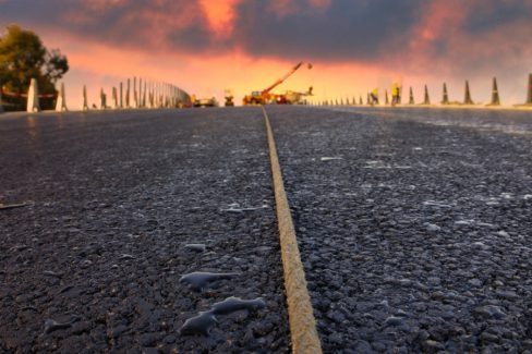 Le soleil se couche sur le nouvel échangeur de la Roche sur Yon. La photographie a été prise lors d'un suivi de chantier en partie nocturne. Le ciel est rouge et violet dans le fond. Au premier plan, une corde, posée sur le bitume encore frais semble enjamber le pont. L'humidité ambiante laisse apparaître des gouttes à côté de la corde. Cette dernière servira à réaliser un pré-marquage des lignes de franchissement.