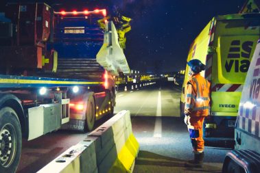 Photographies de nuit prise lors d'un suivi photographique et reportage de chantier nocturne. Un homme en orange de la société Esvia, attend qu'un camion muni d'une grue dépose une BT4- Il s'agit d'une glissière de sécurité destinée à séparer une voie en travaux de la route dite "normale". Pendant toute la nuit, ils vont déposer ces glissières en béton afin de faciliter les travaux de création d'une nouvelle voie en direction de Vannes. Les cônes orange sont de sortie.