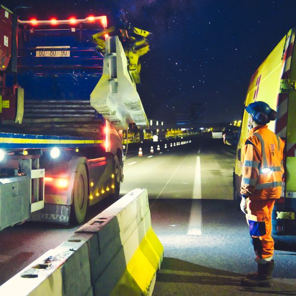 Photographies de nuit prise lors d'un suivi photographique et reportage de chantier nocturne. Un homme en orange de la société Esvia, attend qu'un camion muni d'une grue dépose une BT4- Il s'agit d'une glissière de sécurité destinée à séparer une voie en travaux de la route dite "normale". Pendant toute la nuit, ils vont déposer ces glissières en béton afin de faciliter les travaux de création d'une nouvelle voie en direction de Vannes. Les cônes orange sont de sortie.