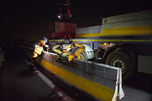 Lors d'un suivi de chantier de nuit, un homme casqué bardé de lampes est en train de fixer une bordure de sécurité à l'aide d'une goupille.