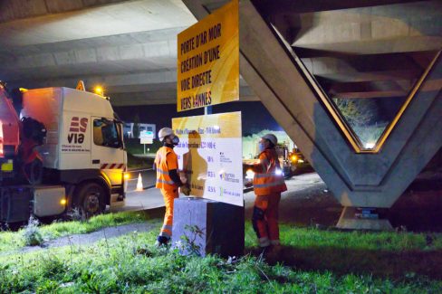 Deux hommes masqués et casqués de la société Esvia sont en train d'installer un panneau dans un giratoire.