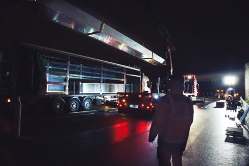 photographies de nuit du déchargement d'un semi-remorque. Sur cette image, un homme casqué surveille l'opération. Une énorme pièce de métal, de type inoxydable, car toutes les lumières se reflètent, est en l'air suspendu au bon vouloir et la dextérité d'un grutier.