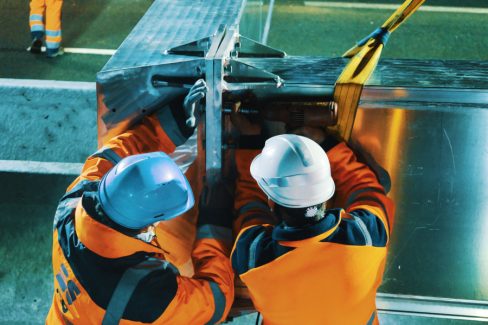 Deux hommes en orange et casqués établissent la jonction de deux segments d'une potence. Ici, dans cette série de photographies de nuit, un des hommes tient une boulonneuse tandis que l'autre fait pression afin de faciliter le serrage des boulons d'un diamètre imposant