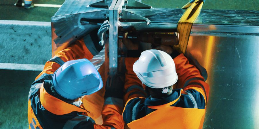 Boulonnage serré Deux hommes en orange et casqués établissent la jonction de deux segments d'une potence. Ici, dans cette série de photographies de nuit, un des hommes tient une boulonneuse tandis que l'autre fait pression afin de faciliter le serrage des boulons d'un diamètre imposant