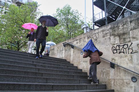 A Paris, au centre Beaubourg, un homme qui semble âgé gravit les marches avec un parapluie cassé posé sur la tête.