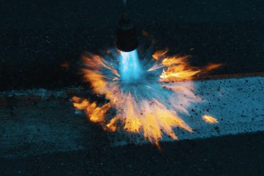 photo prise lors d'un suivi nocturne de chantier. Un ouvrier est en train de passer le chalumeau sur une bande blanche de marquage au sol. L'objectif est d'effacer le marquage temporaire en orange, pour ne laisser place qu'au marquage blanc. Le bleu du gaz qui s'échappe du chalumeau brille de mille feux dans la nuit.