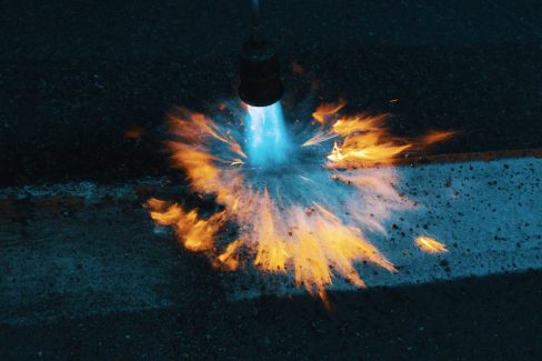 photo prise lors d'un suivi nocturne de chantier. Un ouvrier est en train de passer le chalumeau sur une bande blanche de marquage au sol. L'objectif est d'effacer le marquage temporaire en orange, pour ne laisser place qu'au marquage blanc. Le bleu du gaz qui s'échappe du chalumeau brille de mille feux dans la nuit.