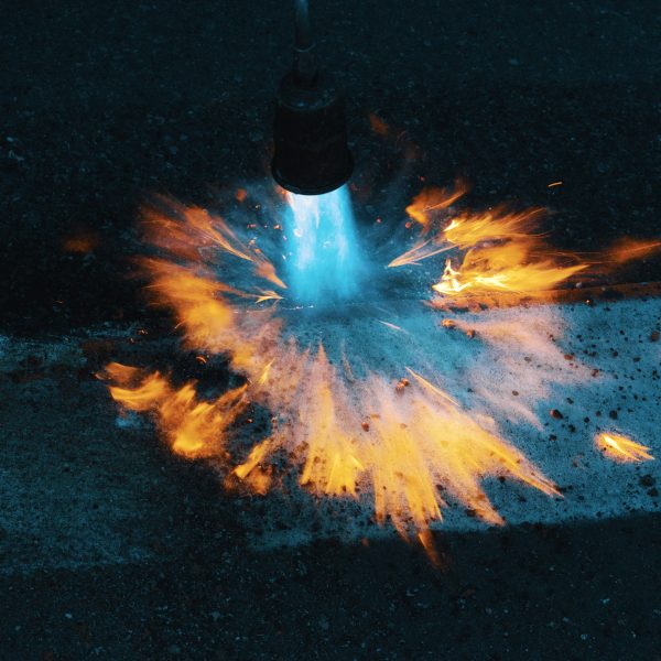 photo prise lors d'un suivi nocturne de chantier. Un ouvrier est en train de passer le chalumeau sur une bande blanche de marquage au sol. L'objectif est d'effacer le marquage temporaire en orange, pour ne laisser place qu'au marquage blanc. Le bleu du gaz qui s'échappe du chalumeau brille de mille feux dans la nuit.
