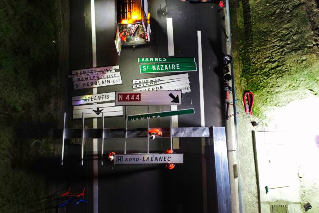 Suivis de chantiers routiers en photos. Des pancartes de signalisations sont en cours de montage sur une potence posée au sol. La prise de vue nocturne par drone est régie par certaines règles quant aux vols par drone.