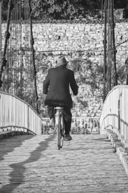 Photo en noir et blanc qui semble datée d'une autre époque. Le cycliste de dos, semble plutôt âgé. Il est vêtu d'un costume et port élégamment un chapeau. Le pont est composé de pavés. Un ponton sans âge.