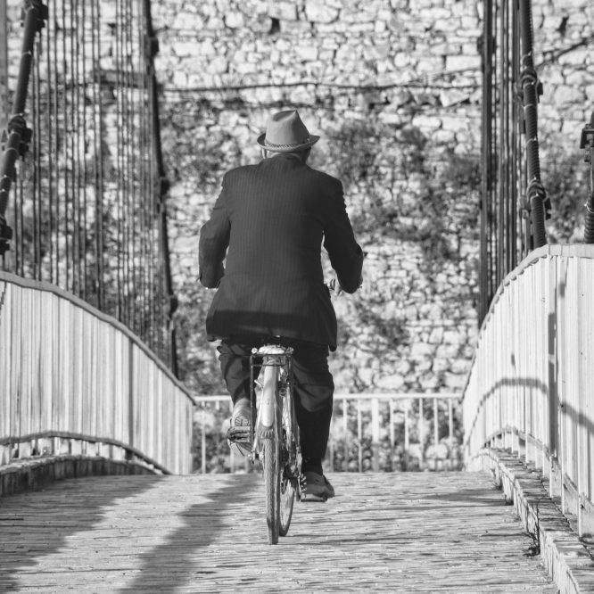 Photo en noir et blanc qui semble datée d'une autre époque. Le cycliste de dos, semble plutôt âgé. Il est vêtu d'un costume et port élégamment un chapeau. Le pont est composé de pavés. Un ponton sans âge.