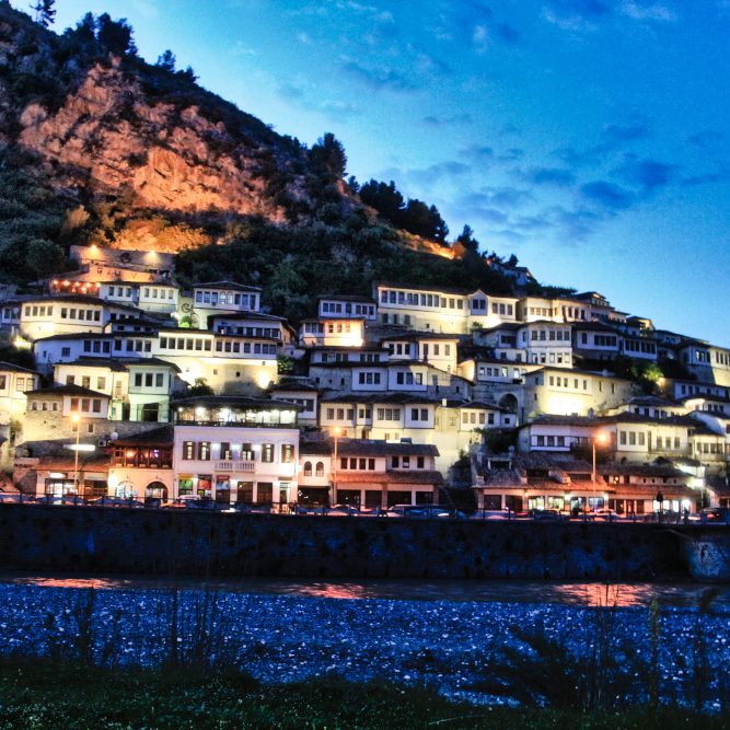 Destination Berrat en Albanie. Un magnifique village qui prend tout son sens et ses couleurs à la nuit tombée. Accrochées à un flanc de montagne les candélabres éclairent les façades blanches aux fenêtres de bois de cette ville quasi antique