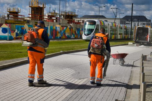 Deux hommes en orange sont équipés de souffleuses. Alors que le tramway très coloré d'Angers s'approche de son arrêt place Terra Botanica, ils soufflent le surplus de gravillons qu'ils viennent de fixer au sol à l'aide d'une résine afin de dessiner un béton matricé.