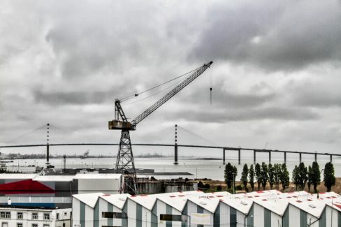 Devant le port de Saint Nazaire, une grue domine la zone industrielle. Le ciel est gris et bas.