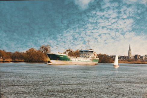 Dans cette photo prise depuis les quais du Pellerin à proximité de Nantes, deux bateaux se livrent une course dont l'issue est déjà connue. UN gracier croise la course d'un voilier qui semble si petit à son côté. Le ciel est rempli de nuages moutonneux et le fier clocher de la ville de Couëron domine la Loire.