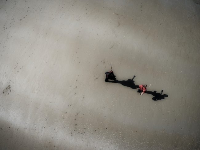 Deux hommes, guitares en bandoulière, sont en train de marcher sur la plage. La photographie est prise au nadir. L'ombre des deux hommes dessinent leurs nobles intentions.