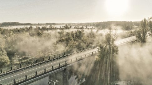Dans cette photographie, aux tons sépias, la brume joue avec les rayons du soleil. Le drone est situé aux abords d'un pont et d'une rivière. Rivière dont s'échappe de magnifiques ondes de brume. Le soleil rasant traversant les arbres vient sectionner cette brume par endroits.