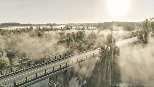 Dans cette photographie, aux tons sépias, la brume joue avec les rayons du soleil. Le drone est situé aux abords d'un pont et d'une rivière. Rivière dont s'échappe de magnifiques ondes de brume. Le soleil rasant traversant les arbres vient sectionner cette brume par endroits.