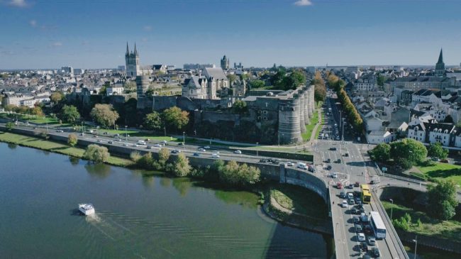 Vue aérienne par drone du château d'Angers. Le pont de la Basse Chaîne est légèrement saturé. Un bateau passe sur le Maine au pied du château. Au fond, sur un ciel servant de fond bleu, on distingue les clochers de la cathédrale.