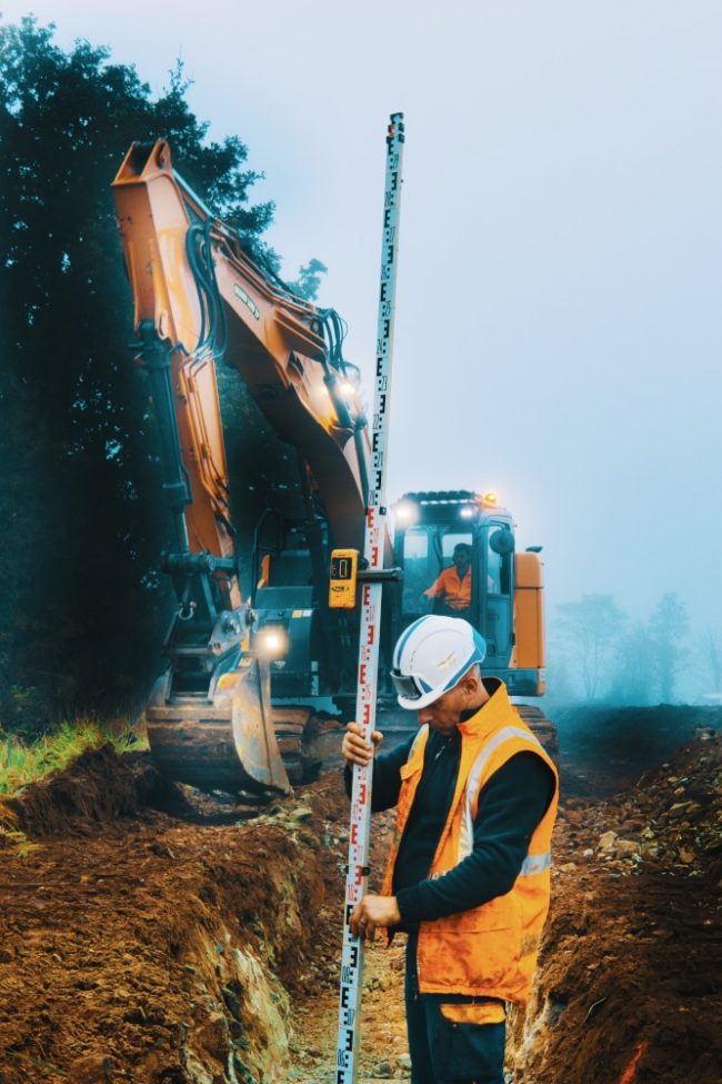 Mesure et tranchée C'est en tant que photographe chantier que nous sommes intervenus sur ce suivi. Un homme en orange et casqué est en train de vérifier à l'aide d'un laser si la tranchée que la pelle en second plan est suffisamment profonde pour accueillir des tuyaux destinés à transporter de l'eau potable.
