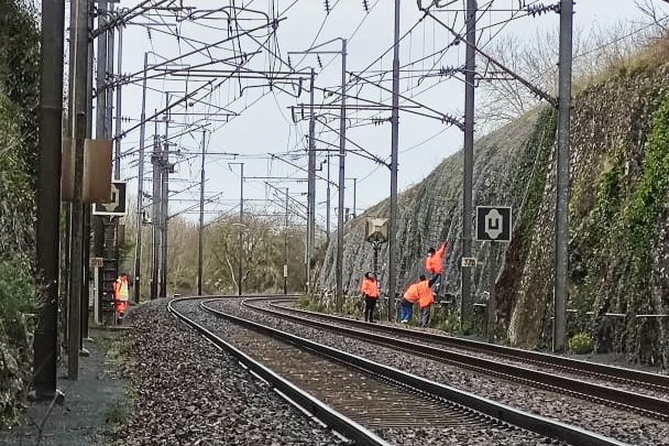 Des hommes vêtus de orange vérifient et inspectent les derniers travaux de renforcement d'une paroi en bordure de voie ferrée. Des prises de vues techniques par drone seront effectuées pour l'établissement du DOE