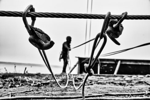 Photographie en noir et blanc lors Dun suivi de chantier. Un système de cordage, de manilles et autres mousquetons sécurisent la descente d'un cage dans laquelle un homme applique du béton projeté.