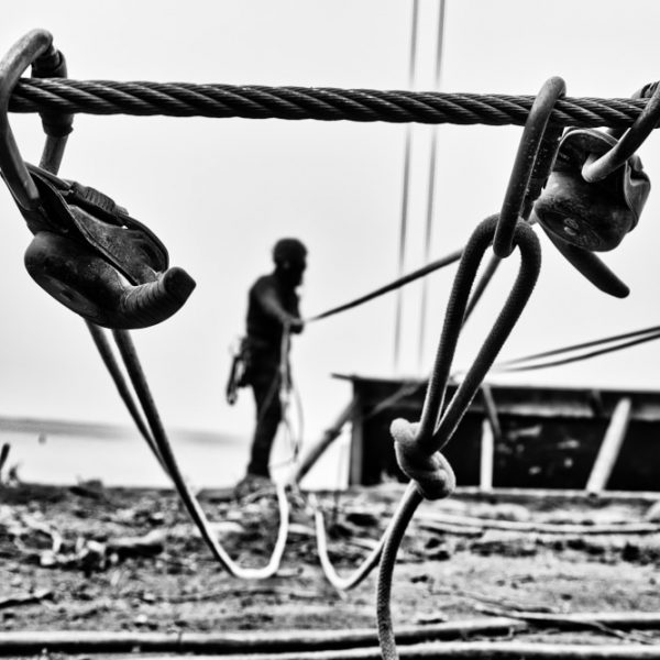 On se rappelle Photographie en noir et blanc lors Dun suivi de chantier. Un système de cordage, de manilles et autres mousquetons sécurisent la descente d'un cage dans laquelle un homme applique du béton projeté.
