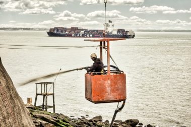 Un homme dans une nacelle applique du béton projeté sur une falaise ayant besoin d'être renforcée. Au large de St Nazaire, un porte-conteneur passe en second plan.