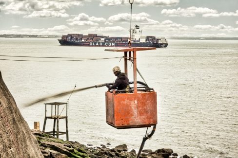Un homme dans une nacelle applique du béton projeté sur une falaise ayant besoin d'être renforcée. Au large de St Nazaire, un porte-conteneur passe en second plan.