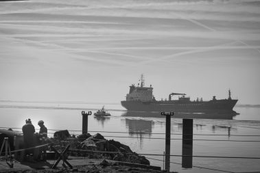 dans le port de St Nazaire, deux ouvriers casqués travaillent au premier plan. Un remorqueur au large passe devant un roulier.