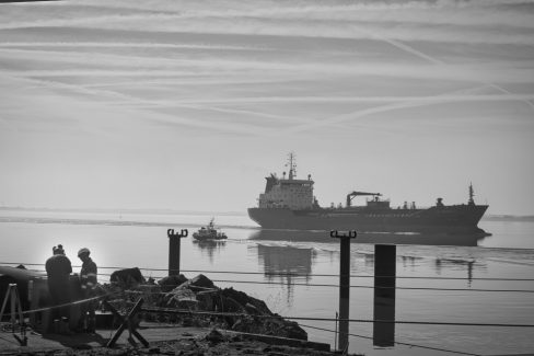 dans le port de St Nazaire, deux ouvriers casqués travaillent au premier plan. Un remorqueur au large passe devant un roulier.