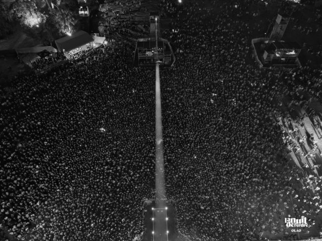 Photographie en noir et blanc prise par drone lors d'un festival. Un faisceau mulineux traverse la foule pour éclairer la sombre scène