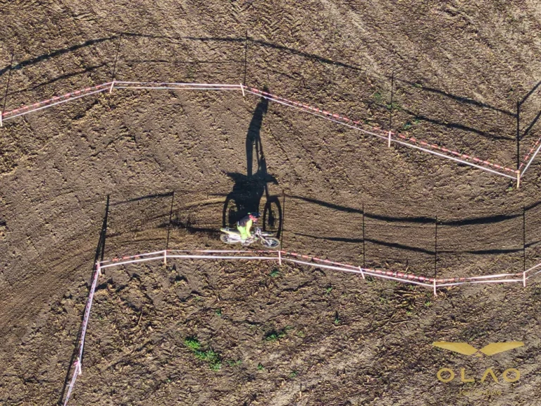 Vue zénithale par drone d'un pilote d'enduro dans un virage balisé, son ombre allongée sur la terre